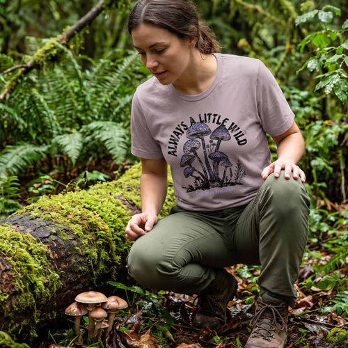 Woman in an Always a Little Wild Pink Gravel tee discovering mushrooms beside a mossy forest log.