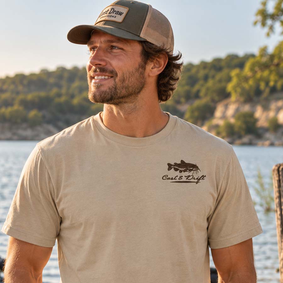 A man wearing a beige short-sleeve t-shirt with a black fish logo and the text 'Cast & Drift' on the left chest, paired with a matching trucker hat, set against a scenic outdoor background of water and trees.