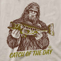 A close-up of a graphic t-shirt featuring a brown line art illustration of Bigfoot holding a green and brown fish, with the text 'CATCH OF THE DAY' printed below in olive green.