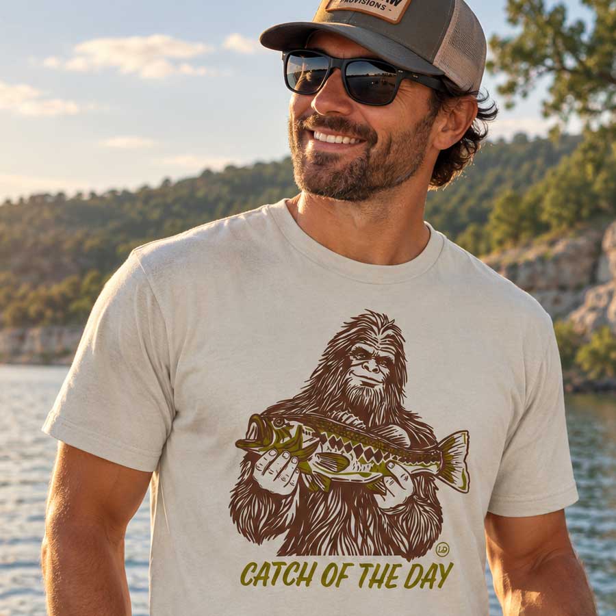 A man wearing a beige graphic t-shirt with a brown and olive-green print of Bigfoot holding a fish and the text 'CATCH OF THE DAY', set against a natural outdoor background of water and trees.