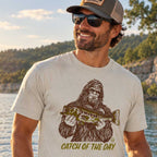 A man wearing a beige graphic t-shirt with a brown and olive-green print of Bigfoot holding a fish and the text 'CATCH OF THE DAY', set against a natural outdoor background of water and trees.