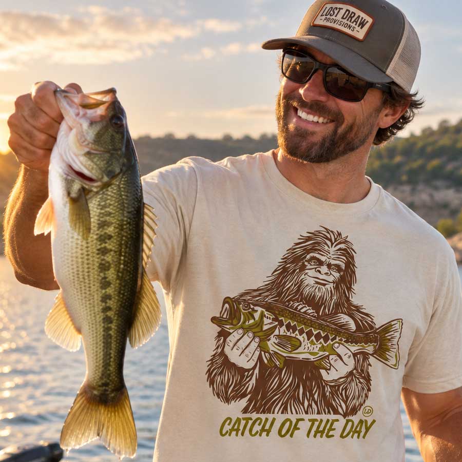 A smiling man wearing a 'Just Draw Provisions' trucker hat and a graphic t-shirt holds up a largemouth bass outdoors by the water at sunset. The t-shirt features a brown graphic of Bigfoot holding a fish with the text 'CATCH OF THE DAY'.