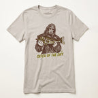 A light beige crewneck t-shirt featuring a graphic of Bigfoot holding a fish, with the text 'CATCH OF THE DAY' printed below in olive green. The brand 'Lost Draw' is visible on the inner neck label.