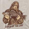 A close-up of a graphic print on a light beige fabric, featuring a brown line art illustration of Bigfoot holding a walleye fish with the text 'CATCH OF THE DAY' underneath.