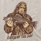 A close-up of a graphic print on a light beige fabric, featuring a brown line art illustration of Bigfoot holding a walleye fish with the text 'CATCH OF THE DAY' underneath.