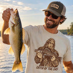 A smiling man outdoors by the water holds up a large fish while wearing a beige graphic T-shirt that reads 'CATCH OF THE DAY' with a large fish illustration, a cap reading 'LOST DRAW', and sunglasses.