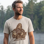 Man wearing the Dust Catch of the Day Walleye Tee on a lakeside dock, with the Bigfoot fishing graphic visible on the front.
