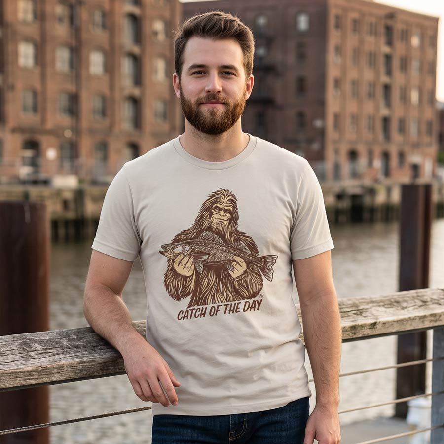 Man wearing a Dust Catch of the Day Walleye Tee beside an urban waterfront, with the Bigfoot fishing design visible.
