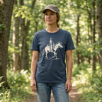Woman wearing the Vintage Navy Dust & Reins Tee on a wooded trail, showing the cream cowgirl and horse graphic.