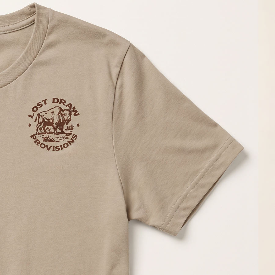 A close-up of a beige short-sleeve t-shirt featuring a brown circular graphic on the left chest with a bison and the text 'LOST DRAW PROVISIONS'.