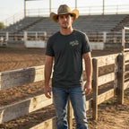 Man wearing the Dark Grey Giddy Up, Y'all Tee at a Texas rodeo arena, showing the small cream-colored front chest graphic.