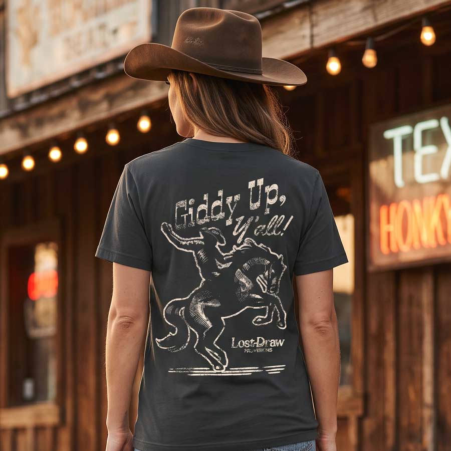 Woman wearing the Giddy Up, Y'all Jersey Tee outside a Texas honky-tonk, displaying the vintage rodeo artwork on the back.