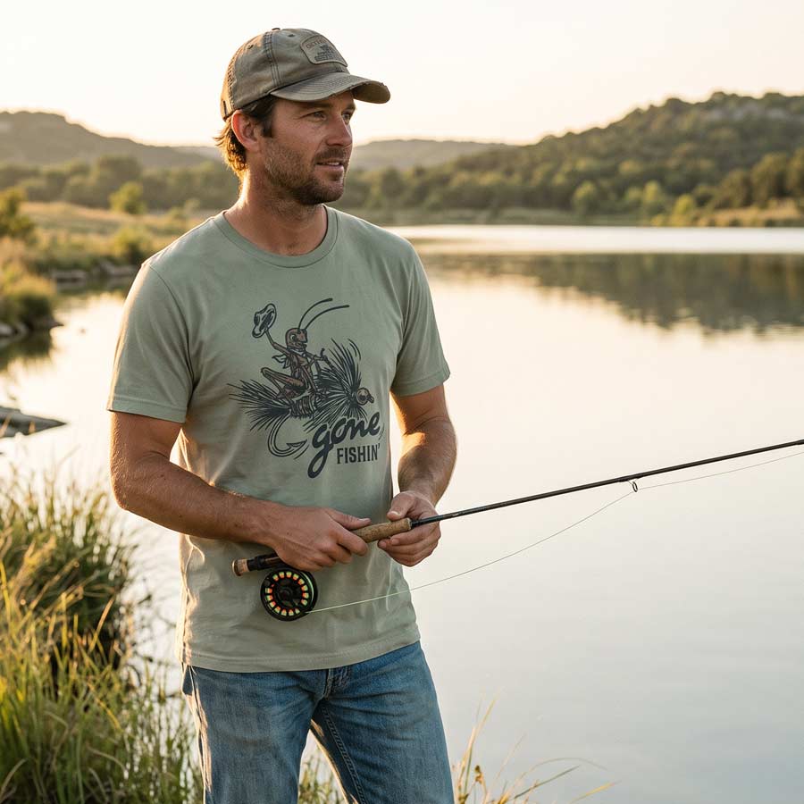 A man stands by a lake at sunset wearing a light sage green t-shirt with a 'Gone Fishing' graphic, blue jeans, and a baseball cap, holding a fly fishing rod.