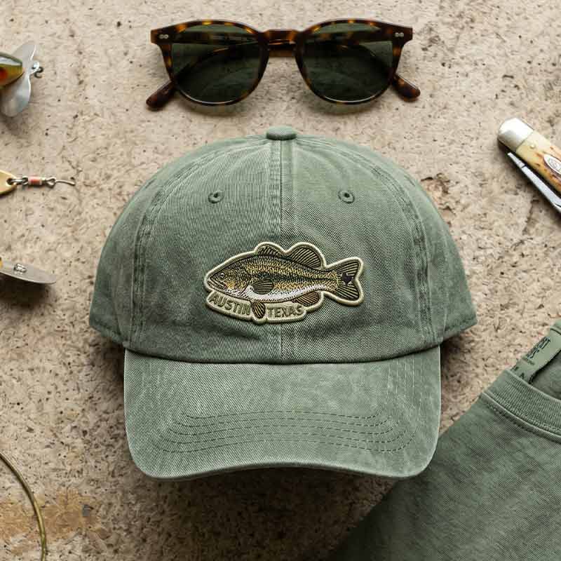 Green cap with fish emblem and 'Austin, Texas' text on a textured surface with sunglasses and knife.