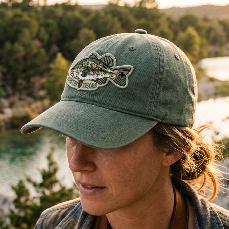 Person wearing a green cap with a fish logo and 'Austin, Texas' text, standing outdoors near water and trees.
