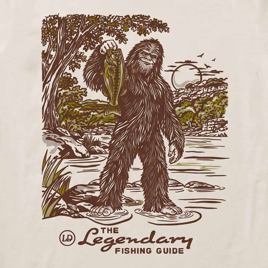 A close-up of a cream-colored fabric surface featuring a vintage-style graphic print of a Bigfoot-like figure holding a fish in a river, with the text 'The Legendary Fishing Guide' and a small 'LD' logo below.