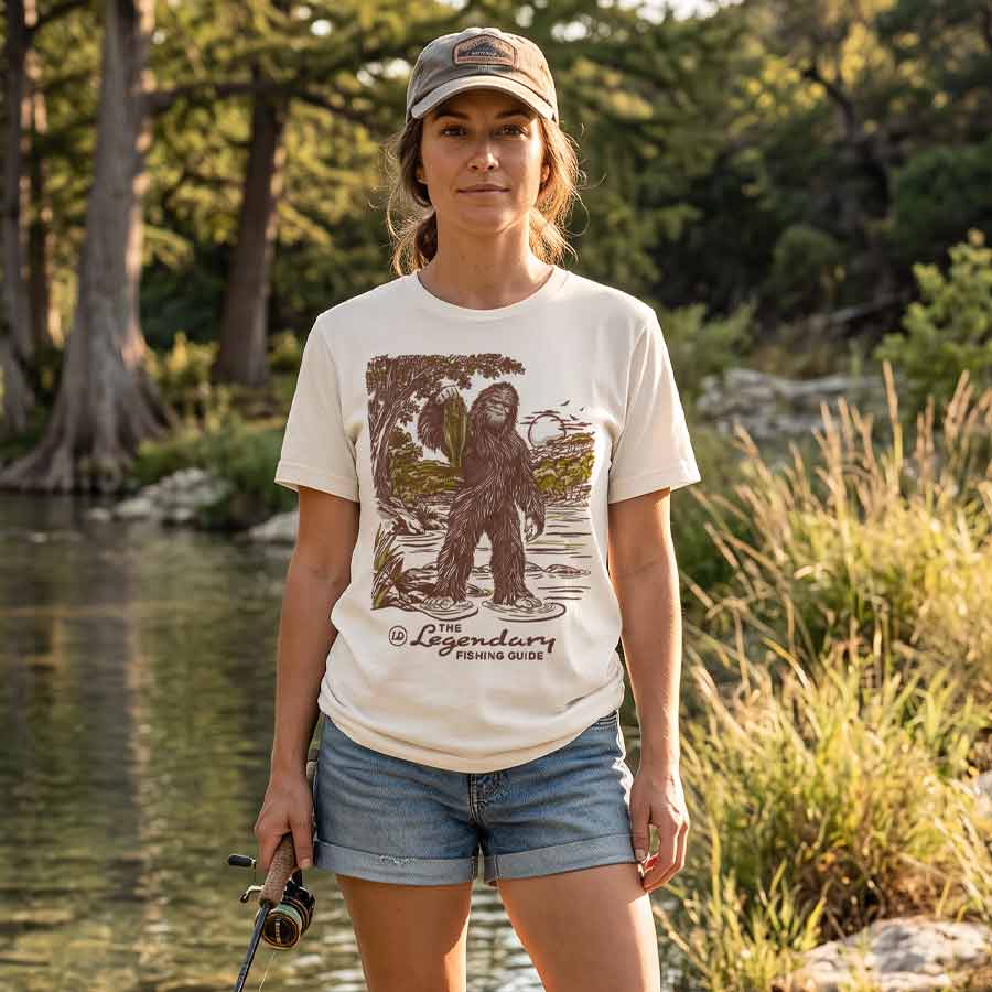 Female model wearing the Lost Draw Provisions Bigfoot fishing graphic tee in Vintage White beside a wooded river.