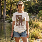 Female model wearing the Lost Draw Provisions Bigfoot fishing graphic tee in Vintage White beside a wooded river.