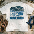 Heavyweight oversized white graphic tee Blue Hole Georgetown TX on a stone surface with sandals and a bandana.