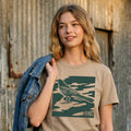 Woman wearing khaki garment-dyed City Camo Austin boxy t-shirt with bird graphic in front of a rustic wooden wall.