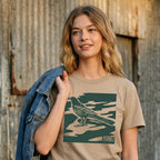 Woman wearing khaki garment-dyed City Camo Austin boxy t-shirt with bird graphic in front of a rustic wooden wall.