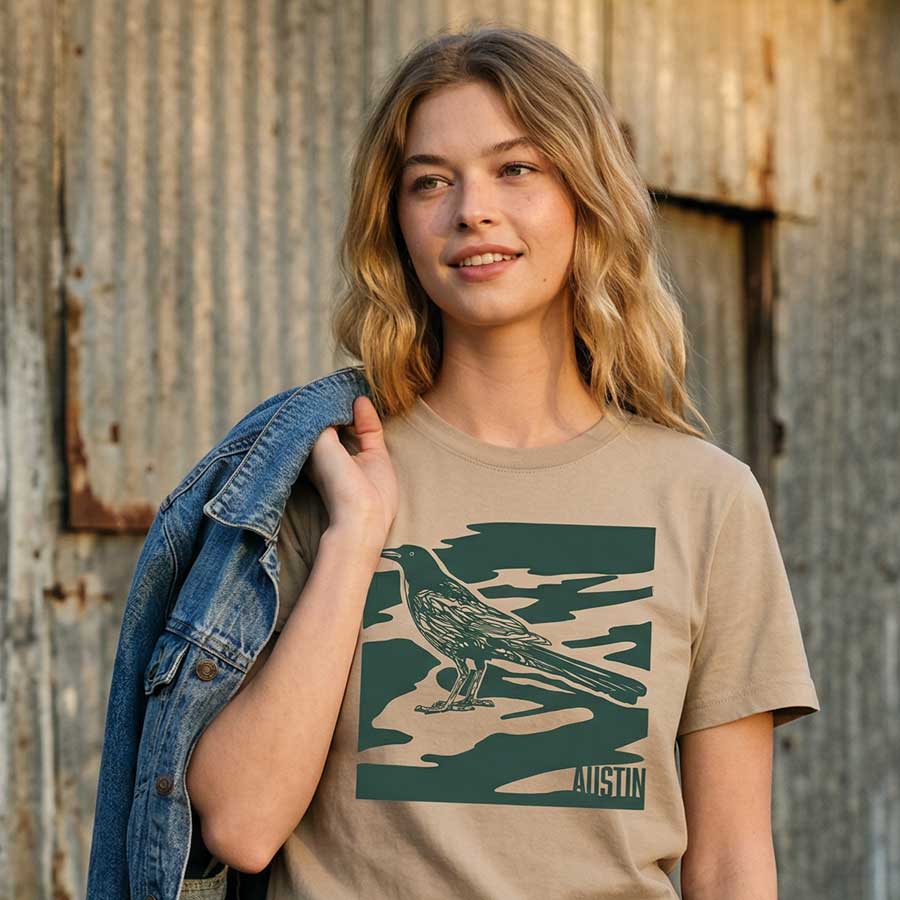 Woman wearing khaki garment-dyed City Camo Austin boxy t-shirt with bird graphic in front of a rustic wooden wall.