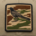 Embroidered patch of a bird on a camouflage background on a beige cap.