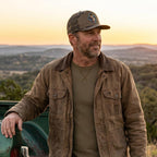 Man standing outdoors with a scenic background, wearing a brown jacket and cap.