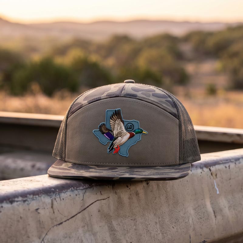 Brown cap with a Texas-shaped duck design on a metal railing with a scenic background