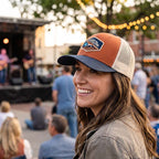 Woman wearing a baseball cap with a patch, smiling at an outdoor event.