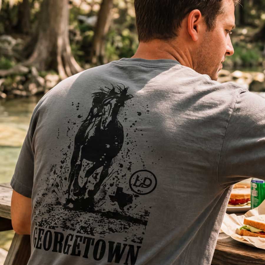 Man wearing Georgetown TX horse graphic tee oversized heavyweight t-shirt western style, sitting outdoors by a body of water.