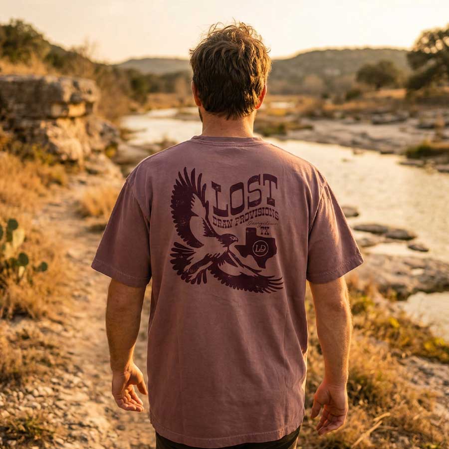 Person wearing a 'Lost Draw Provisions' t-shirt with an hawk design, standing in a scenic outdoor setting.