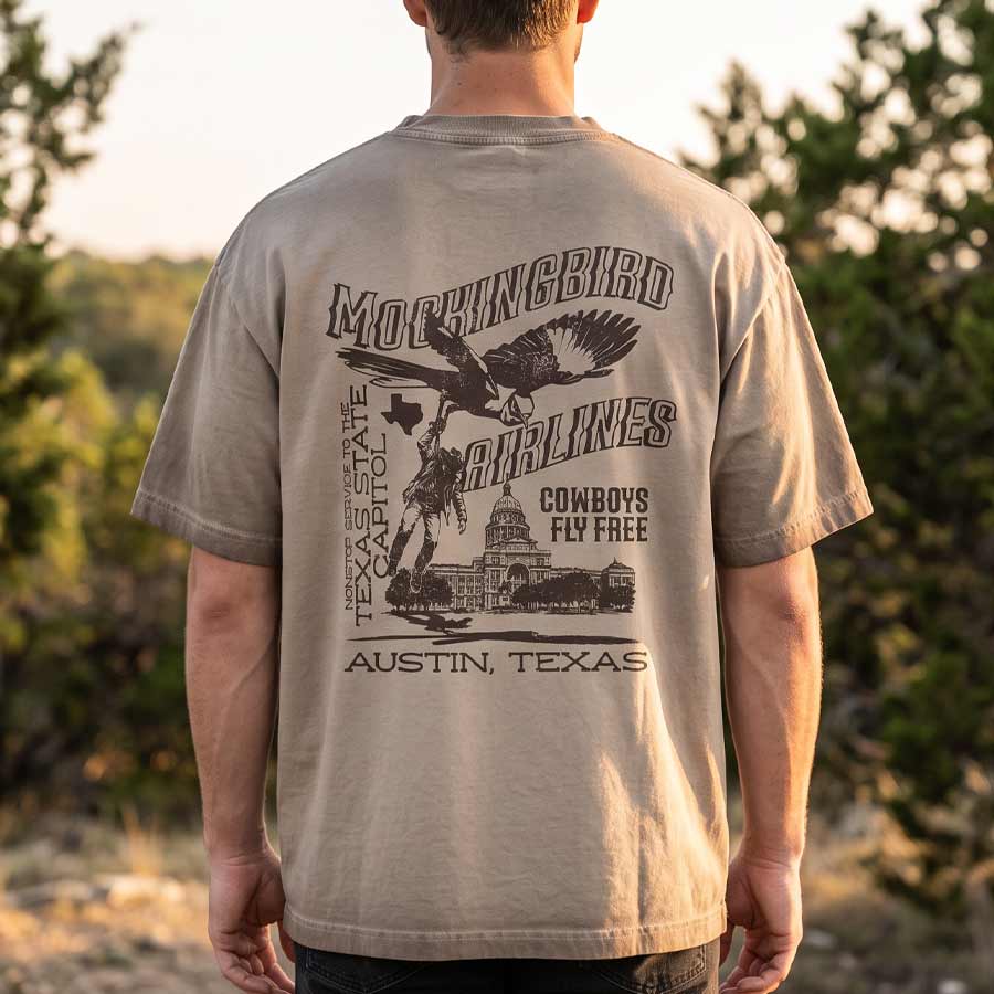 A man viewed from behind wearing a beige t-shirt with a large black graphic print on the back featuring an eagle, cowboy, and the text 'MORNINGBIRD AIRLINES' and 'COWBOYS FLY FREE AUSTIN, TEXAS'.