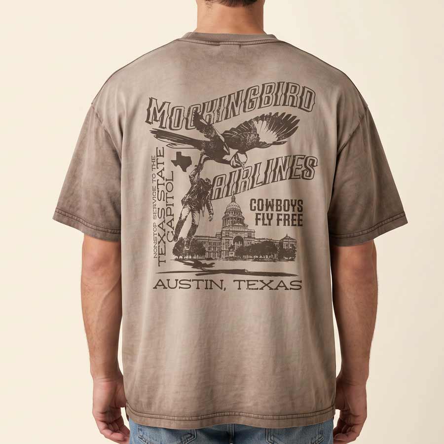 A man wearing a faded brown t-shirt, viewed from the back. The shirt features a large graphic print with the words 'MOCKINGBIRD AIRLINES', 'TEXAS STATE', 'AUSTIN, TEXAS', and 'COWBOYS FLY FREE' alongside an eagle and cowboy illustration.