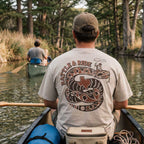 Man in a canoe on a river wearing Rattle and Ride Texas graphic tee oversized tan heavyweight t-shirt Lost Draw Provisions flat lay back