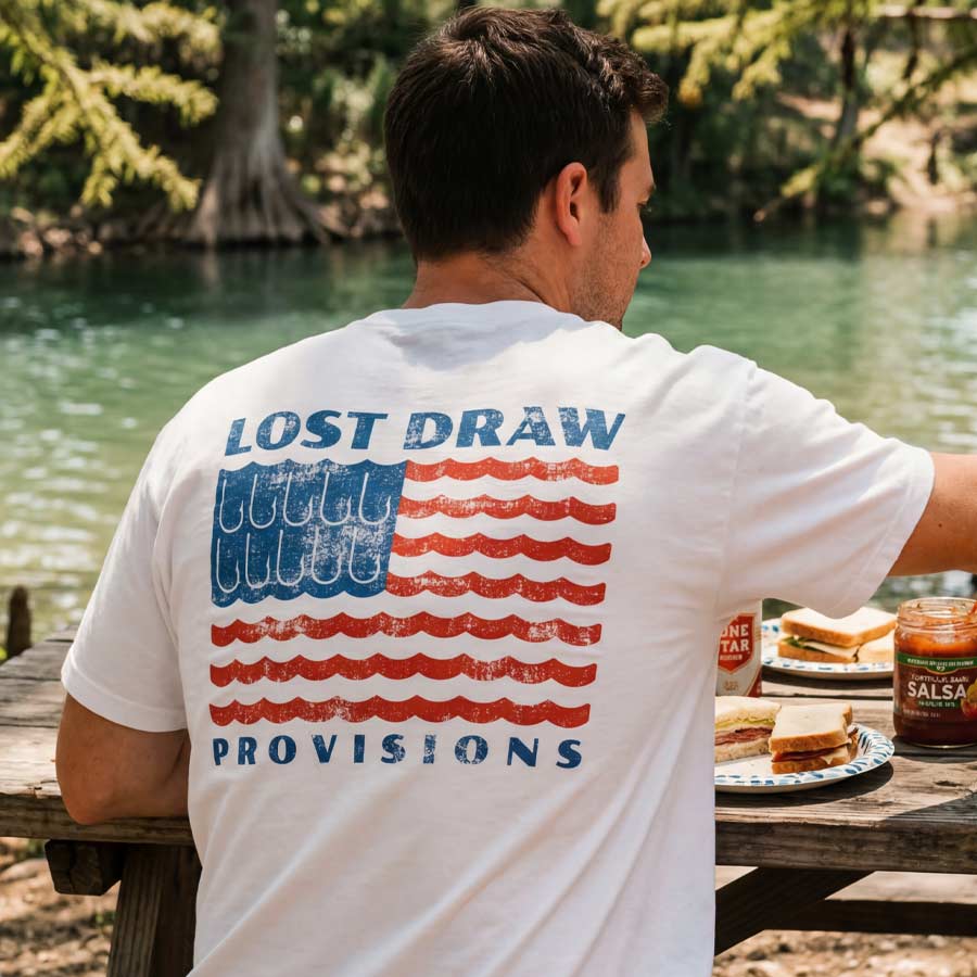 lifestyle image of man wearing American flag fishing hook t-shirt river wave graphic oversized cotton tee