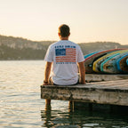 Person sitting on a dock wearing American flag fishing hook t-shirt river wave graphic oversized cotton tee