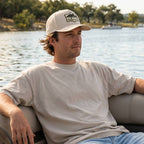 Man sitting by a lake wearing The Blue Hole Bass hat, with trees and water in the background.