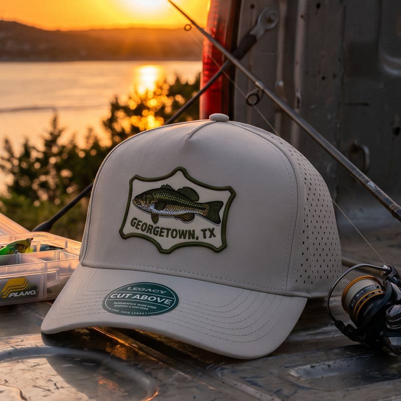 White cap with fish logo and 'Georgetown, TX' text on a fishing rod and tackle box with sunset in the background.