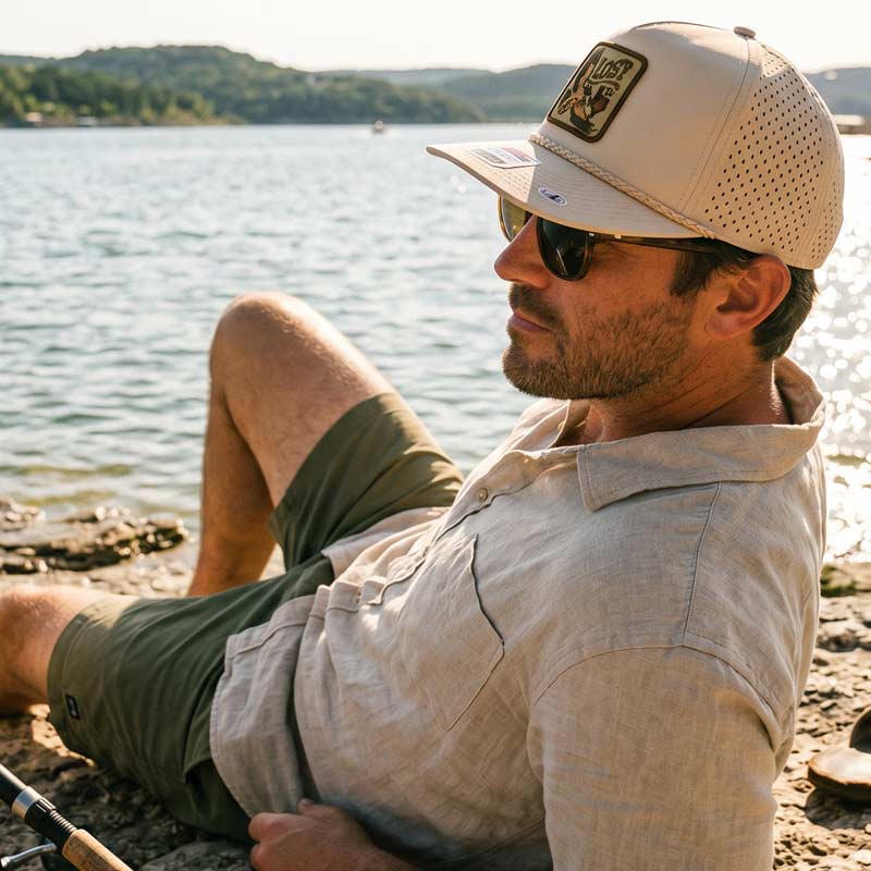 Man sitting by a lake wearing a beige cap and sunglasses, with fishing gear nearby.