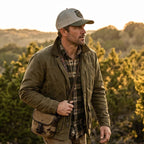 Man in outdoor setting wearing a cap, jacket, and plaid shirt with a bag.