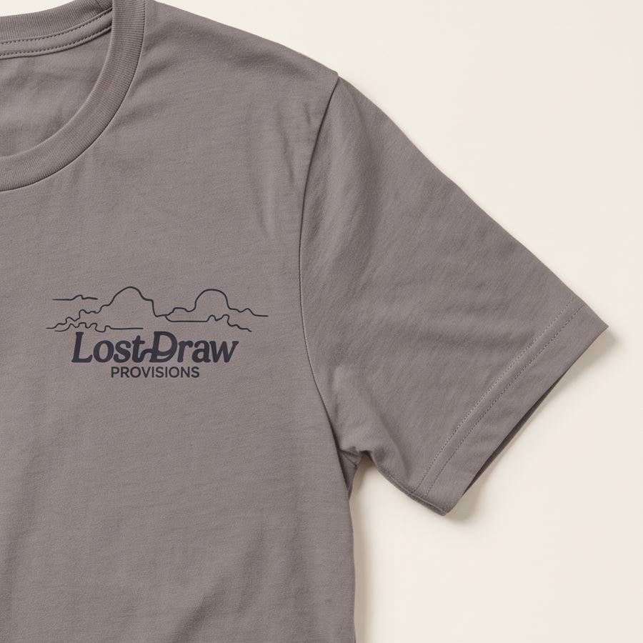A close-up of a pebble brown short-sleeve t-shirt featuring a dark gray printed graphic on the left chest with the text 'Lost-Draw PROVISIONS' and a simple mountain outline, set against a light cream background.