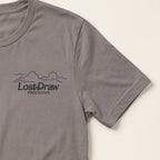 A close-up of a pebble brown short-sleeve t-shirt featuring a dark gray printed graphic on the left chest with the text 'Lost-Draw PROVISIONS' and a simple mountain outline, set against a light cream background.