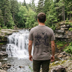 Man wearing the North of Ordinary Jersey Tee beside a waterfall, displaying the vintage outdoor graphic on the back.