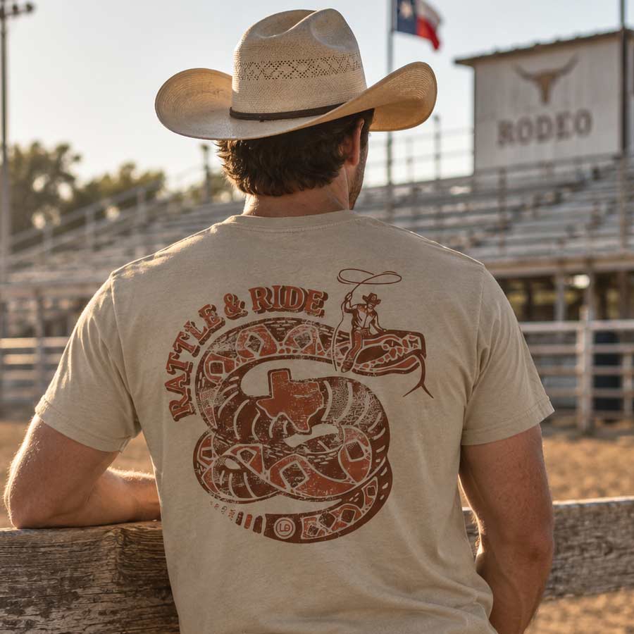 Man wearing the tan Rattle & Ride Tee at a rodeo arena, showing the Rattle and Ride back artwork from Lost Draw Provisions.