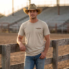 Man wearing the Dark Grey Rattle & Ride Tee at a rodeo arena, showing the small Lost Draw Provisions front chest logo.