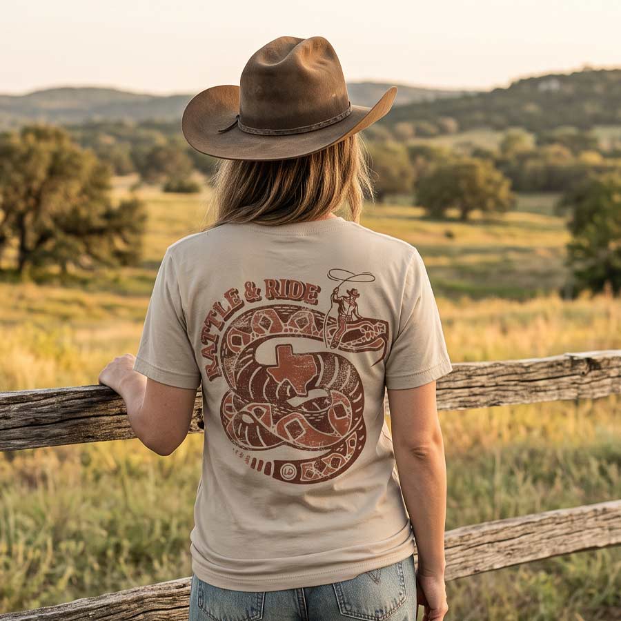 Woman wearing the Rattle & Ride Jersey Tee beside a ranch fence, displaying the vintage rattlesnake and cowboy back artwork.