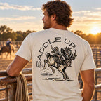 A man wearing a cream-colored t-shirt with a large black graphic on the back, standing in a ranch paddock at sunset. The graphic features a bucking horse with a rabbit on its neck and the text 'SADDLE UP' and 'LostDraw Ranch'.