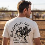 Man wearing the Vintage White Saddle Up Jersey Tee with a cowboy and jackalope back graphic at a Texas rodeo.