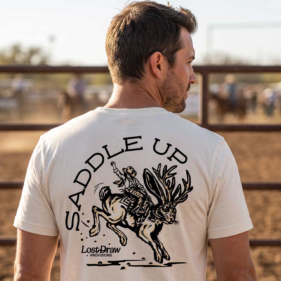 Man wearing the Vintage White Saddle Up Jersey Tee with a cowboy and jackalope back graphic at a Texas rodeo.